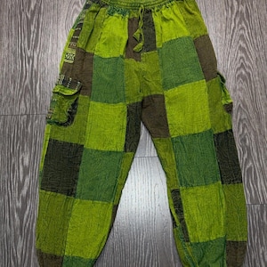 May include: Patchwork pants featuring a checkered pattern in various shades of lime green and dark olive green. The pants have an elastic waistband with a drawstring, side cargo pockets, and elasticized cuffs at the bottom of each pant leg.