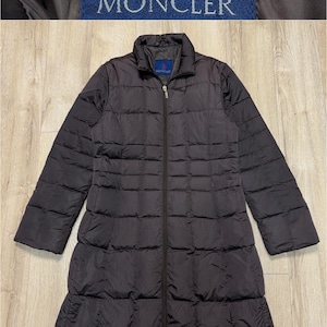 Puede incluir: Un abrigo largo acolchado de color marrón oscuro con cremallera completa. El abrigo tiene cuello y la marca "MONCLER" en un parche azul marino. Diseñado para abrigar y proteger de los elementos.