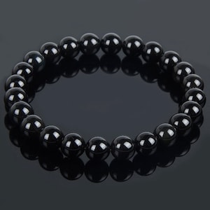 May include: A black beaded bracelet with a glossy finish. The bracelet is made of round, polished beads strung together. The beads reflect light, creating a shiny appearance. The bracelet is displayed on a dark surface.