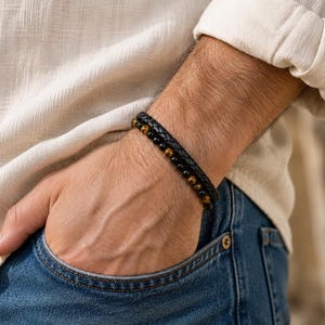 May include: A layered bracelet featuring a braided black leather cord alongside a strand of smooth, round black beads and brown tiger eye gemstone beads. The bracelet is worn on a wrist, showcasing the contrast between the dark leather and the natural stone textures.