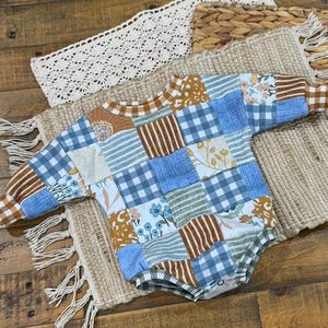 May include: A patchwork baby romper with long sleeves. The romper features a mix of blue gingham, floral, and striped fabric squares. The sleeves and neckline are trimmed in a solid brown fabric. The romper is lying on a woven mat.