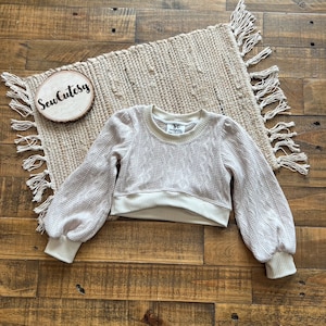 May include: A cream-colored cropped sweater with long sleeves and a cable knit pattern. The sweater has a round neckline and a ribbed hem. The sweater is laying flat on a light brown wooden surface.