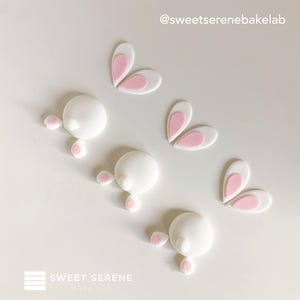 May include: Three sets of edible fondant bunny decorations, each featuring a round white tail, two small pink-padded feet, and two white ears with pink centers. The text @sweetserenebakelab and SWEET SERENE BAKE LAB is visible in the image.