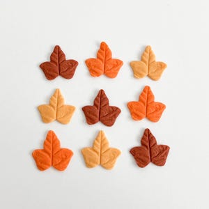 May include: A collection of nine small, flat leaf-shaped decorations arranged in a three-by-three grid. Each piece features a textured surface with a central vein and is colored in various shades of brown, orange, and yellow, resembling autumn foliage.