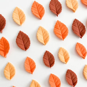 May include: A collection of small, individual leaf-shaped decorations in shades of yellow, orange, and brown. Each piece features a central vein and textured surface, arranged in a scattered pattern across a plain white background.