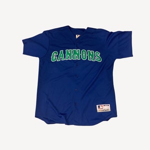 Puede incluir: Una camiseta de béisbol azul con la palabra "CANNONS" en letras verdes en la parte delantera. La camiseta tiene un frente abotonado y un logotipo de la Major League Baseball en la manga derecha. La camiseta está hecha por Majestic.