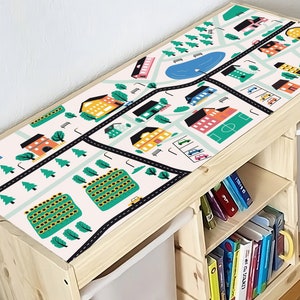 May include: A wooden toy storage bench with a colorful town map design on top. The bench has two white storage bins and a shelf filled with books. The map features roads, buildings, a lake, and trees, creating a playful and educational piece.