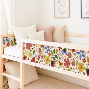May include: A light wood bunk bed with a colorful floral pattern on the headboard and side panels. The pattern includes red, pink, yellow, and blue flowers with green leaves and butterflies. The room has white walls and decorative framed art.