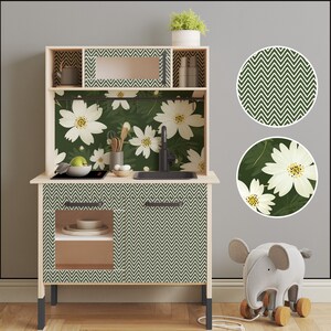 May include: A white toy kitchen playset with green and white floral wallpaper and a chevron pattern on the cabinet doors. The kitchen has a sink, a stovetop, and a shelf. There is a small toy elephant in the foreground.