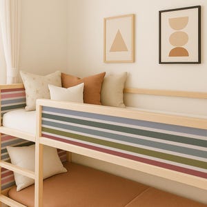May include: A light-coloured wooden bunk bed featuring horizontal striped panels in muted tones of blue, green, pink, and grey. The bed is dressed with a solid brown mattress and several decorative pillows in cream, tan, and star-patterned fabric.