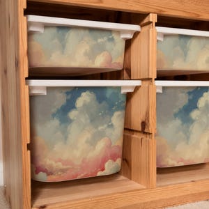 May include: A light-colored wooden storage unit featuring four rectangular plastic bins. Each bin is decorated with a printed graphic of a blue sky filled with white and pink clouds. The unit has a vertical divider separating the two columns of bins.
