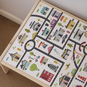 May include: A rectangular wooden table featuring a white tabletop surface printed with a colorful city map graphic. The design includes black roads, various buildings labeled as hospital and police, trees, vehicles, and a hot air balloon illustration.