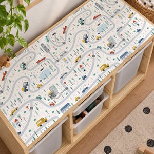 May include: A light-colored wooden toy storage bench featuring a top surface printed with a colorful city street map pattern. The design includes various vehicles, buildings, trees, and road signs in shades of blue, red, yellow, and green.