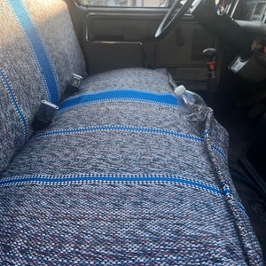 May include: A woven bench seat cover featuring a textured gray and blue pattern with horizontal stripes. The cover is fitted over a vehicle bench seat, showing seat belt buckles and a clear plastic water bottle resting on the textured fabric surface.
