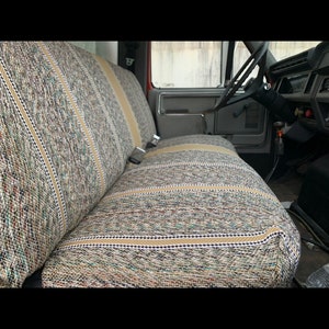 May include: A truck interior featuring a bench seat with a multi-colored, patterned fabric. The seat has a beige stripe down the middle and is secured with a seatbelt. The steering wheel and dashboard are visible.