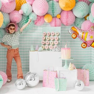 May include: A child poses in front of a party scene with pastel decorations, including honeycomb balls and a roller skate balloon. The scene includes a cake, donuts, gift bags, and disco balls, creating a festive atmosphere.
