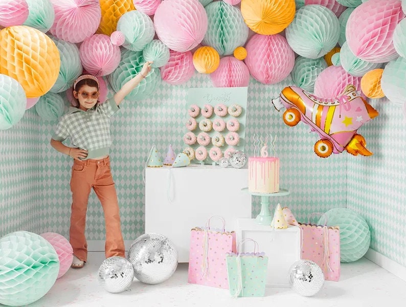 May include: A child poses in front of a party scene with pastel decorations, including honeycomb balls and a roller skate balloon. The scene includes a cake, donuts, gift bags, and disco balls, creating a festive atmosphere.