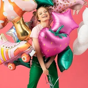 May include: A person holding a collection of colorful foil balloons against a pink background. The balloons include a pink strawberry, green star, and a pink unicorn. Other balloons are shaped like roller skates and stars.