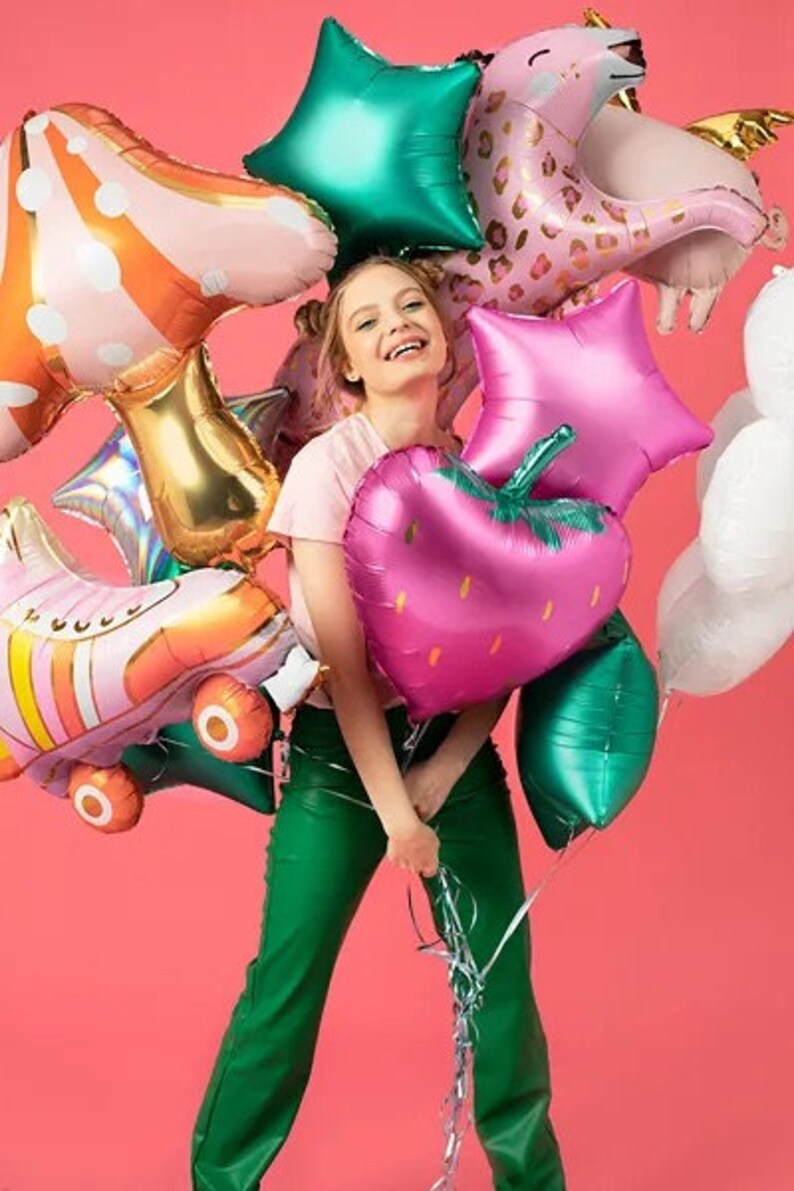 May include: A person holding a collection of colorful foil balloons against a pink background. The balloons include a pink strawberry, green star, and a pink unicorn. Other balloons are shaped like roller skates and stars.