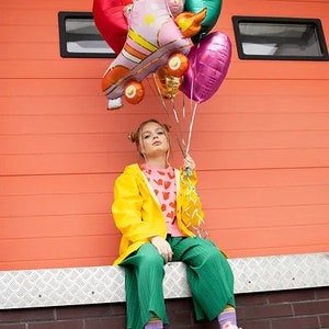 May include: A person wearing pink roller skates, green pants, and a yellow jacket holds a collection of colorful balloons. The balloons include a roller skate design, a heart, and a leaf shape. The background is an orange wall.