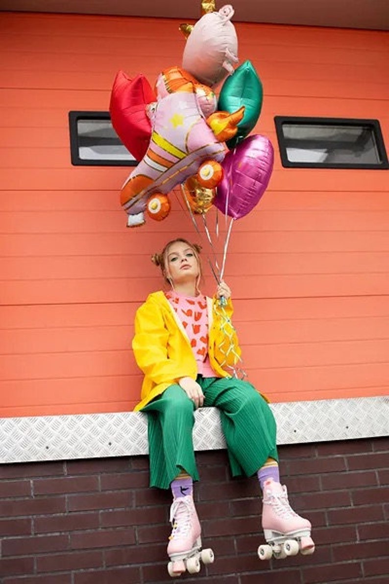 May include: A person wearing pink roller skates, green pants, and a yellow jacket holds a collection of colorful balloons. The balloons include a roller skate design, a heart, and a leaf shape. The background is an orange wall.