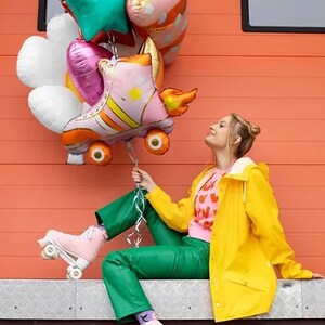 May include: A collection of colorful balloons, including a roller skate design, held by a person wearing pink roller skates and green pants. The person is wearing a yellow jacket and sitting in front of an orange wall.