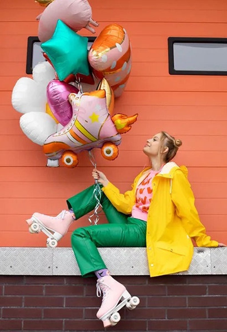 May include: A collection of colorful balloons, including a roller skate design, held by a person wearing pink roller skates and green pants. The person is wearing a yellow jacket and sitting in front of an orange wall.