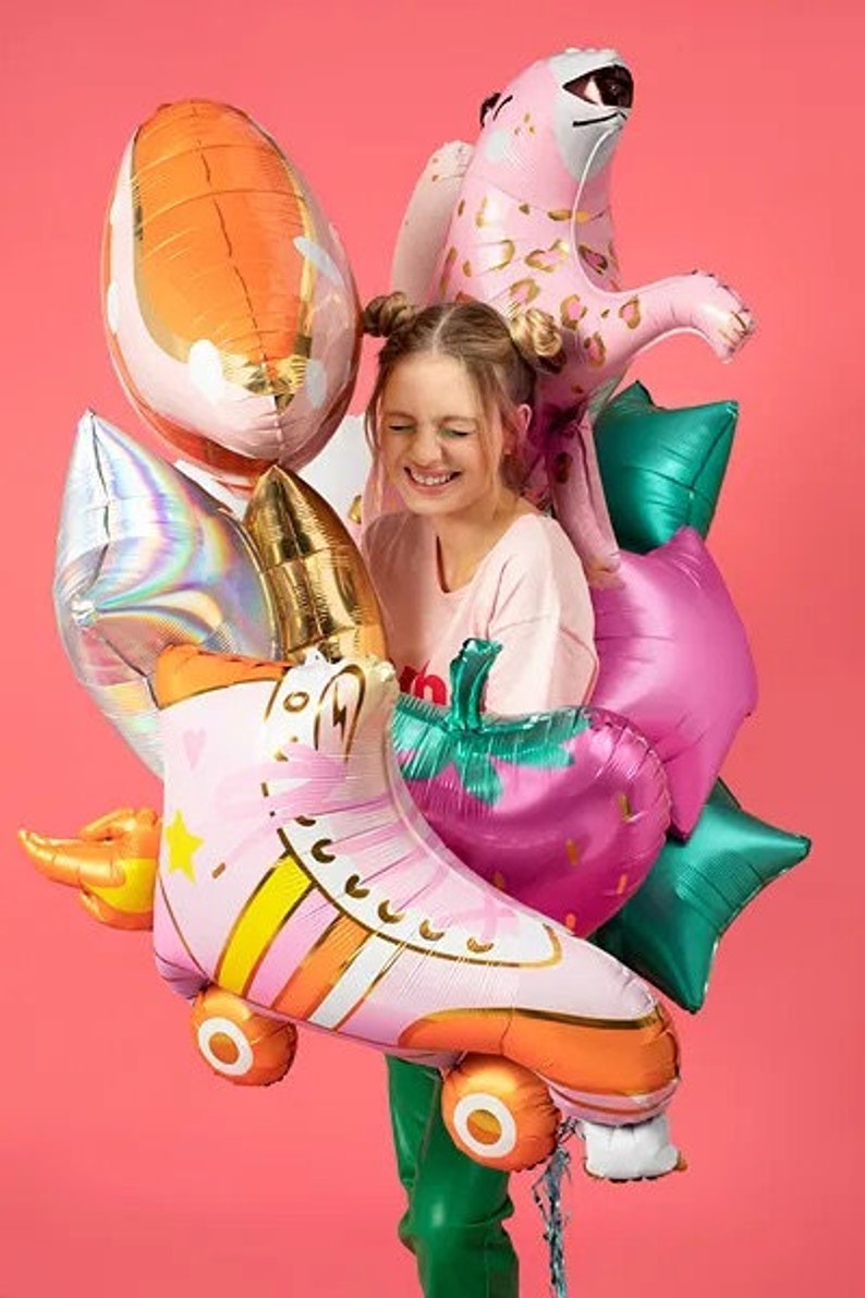 May include: A collection of colorful foil balloons, including a pink roller skate, a pink leopard, a strawberry, and star-shaped balloons. The balloons are in shades of pink, orange, green, and gold, creating a festive and playful atmosphere.