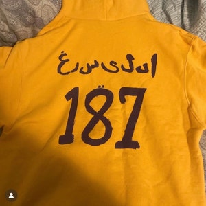 May include: Yellow hoodie with a purple Arabic phrase and the number 187 printed on the back.