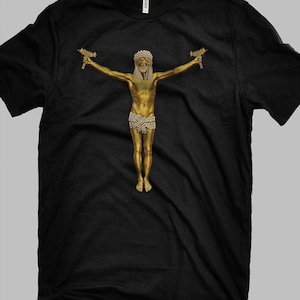 Może przedstawiać: Czarny t-shirt z pozłacaną statuą postaci z wyciągniętymi ramionami, trzymającej dwa zabawki-pistolety. Postać jest ozdobiona koroną i wysadzanym klejnotami przepaską na biodrach. Koszulka jest wykonana z miękkiego materiału.