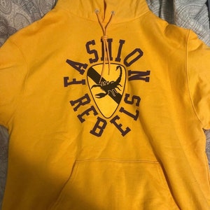May include: A yellow hoodie with a purple graphic of a scorpion inside a shield with the text "FASHION REBELS" around it.