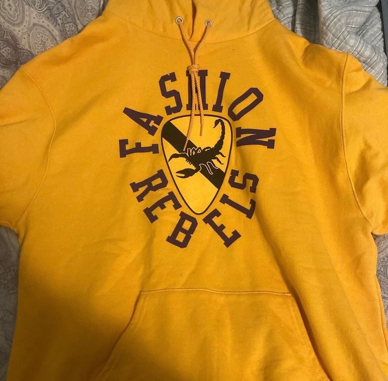 May include: A yellow hoodie with a purple graphic of a scorpion inside a shield with the text "FASHION REBELS" around it.