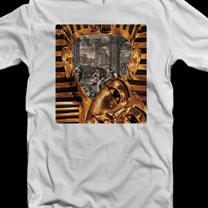 Op de afbeelding: Een wit T-shirt met een gouden en zwarte afbeelding van een farao's masker. Het masker wordt gedeeltelijk bedekt door een scène met mensen in een stad.
