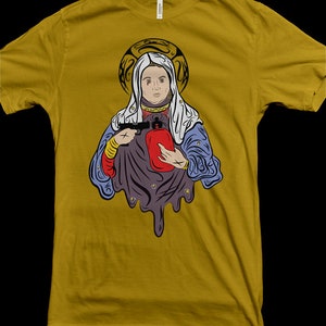 May include: A mustard yellow t-shirt featuring a graphic print of a person holding a red object and a gun. The person is wearing a blue and white robe with a halo above their head.