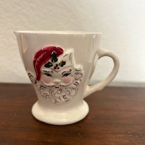 May include: A white ceramic mug with a raised Santa Claus face design. The Santa has a red hat, a white beard, and a pink face. The mug has a gold rim and a handle.