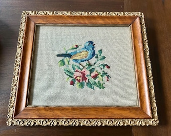 May include: A framed needlepoint embroidery featuring a blue bird with yellow accents perched on a branch with pink flowers. The frame is made of wood with a gold-colored ornate border.