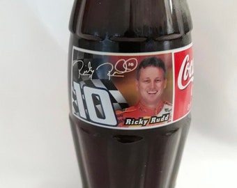 May include: A glass Coca-Cola bottle with a black label featuring a photo of race car driver Ricky Rudd and the number 10. The label also includes the Coca-Cola logo and Rudd's signature.