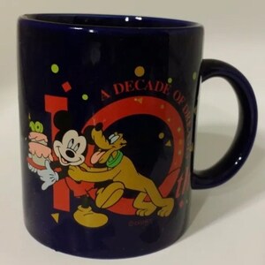 Puede incluir: Una taza de café de cerámica azul con una imagen de Mickey Mouse y Pluto celebrando una década de sueños. La taza tiene un 10 rojo y amarillo con el texto "A Decade of Dreams" alrededor.