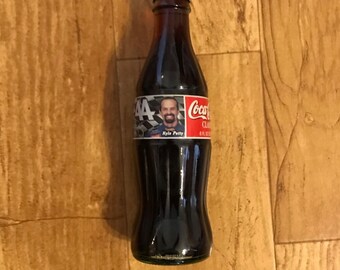 May include: A glass Coca-Cola bottle with a red and white label featuring a photo of race car driver Kyle Petty. The label also includes the number 44 and the text "Coca-Cola Classic".