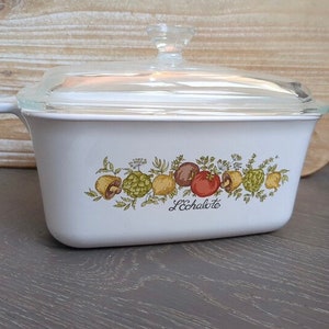 Puede incluir: Plato de cerámica blanco para hornear con tapa de vidrio. El plato tiene un diseño floral con verduras y el texto "L'Echalote".