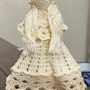 Könnte beinhalten: Eine gehäkelte Puppe mit langen weißen Haaren, einem weißen Kleid und einer weißen Krone. Die Puppe steht auf einem weißen Sockel.
