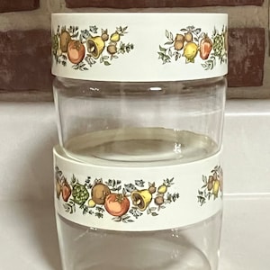 Könnte beinhalten: Zwei durchsichtige Glasbehälter mit weißen Deckeln und einem Blumendesign mit Früchten, Gemüse und Glocken. Das Design ist in Grün-, Gelb-, Orange- und Brauntönen gehalten.