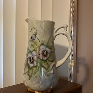 May include: A vintage ceramic pitcher with a bulbous base and a curved handle. The pitcher is painted with purple and white floral designs and green leaves. The pitcher has a gold-colored base.