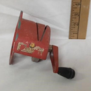 Pode incluir: Um descascador de maçãs vintage vermelho de metal com manivela e cabo preto. O descascador tem uma base circular e uma caixa de lâmina com uma abertura em forma de V. Uma régua de madeira está no fundo.