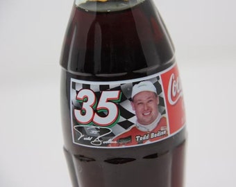 May include: A Coca-Cola bottle with a label featuring NASCAR driver Todd Bodine. The label has a checkered flag background, the number 35, and Bodine's signature.