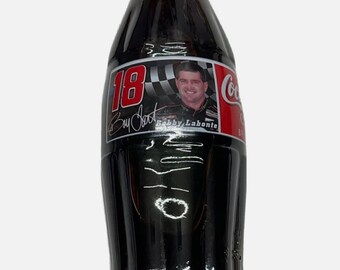 May include: A black glass Coca-Cola bottle with a silver cap. The bottle has a label with a picture of a race car driver, the number 18, and the text "Bobby Labonte".