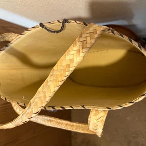 K&ouml;nnte beinhalten: Eine geflochtene Strohtasche mit schwarzem Besatz und einem einzigen geflochtenen Henkel. Die Tasche ist leer und das Innere ist sichtbar.