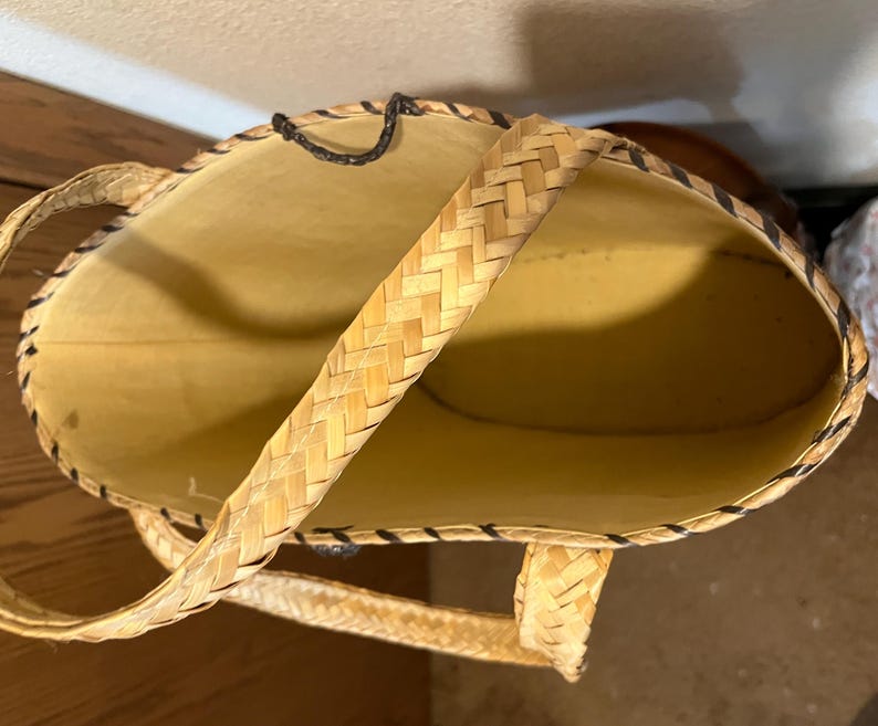 K&ouml;nnte beinhalten: Eine geflochtene Strohtasche mit schwarzem Besatz und einem einzigen geflochtenen Henkel. Die Tasche ist leer und das Innere ist sichtbar.