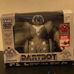 Op de afbeelding: Een grijs en wit robot speelgoed met een rode dart shooter. De doos zegt "Dartbot" en heeft een Bluetooth logo. De robot heeft een wit gezicht met een zwart oog en een grijze helm. De doos zegt ook "Leeftijd 10+" en "Snelle schietdart".