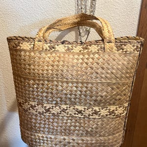 K&ouml;nnte beinhalten: Eine gro&szlig;e, gewebte Tragetasche aus nat&uuml;rlichen Materialien. Die Tasche hat eine rechteckige Form mit einer breiten Basis und einem schmalen oberen Teil. Die Tasche ist aus einem hellbraunen Material mit einem gewebten Muster gefertigt. Die Tasche hat zwei Henkel, die in die Tasche eingewebt sind.